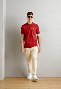 Model in einem enganliegenden roten Polohemd mit Kragen, hellbeigen Hosen und weißen Sneakern, vor einem hellgrauen Hintergrund.