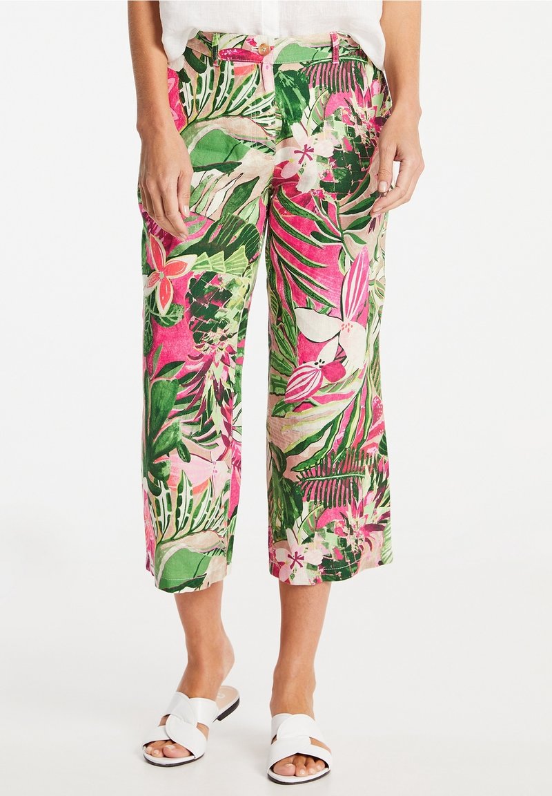 Femme portant un pantalon court imprimé floral tropical rose, vert et blanc et des sandales blanches à enfiler sur fond blanc.