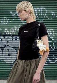 Camiseta preta com logótipo, saia plissada verde-oliva e uma mala preta com um brinquedo de peluche castanho claro. O fundo mostra graffiti verde e branco.