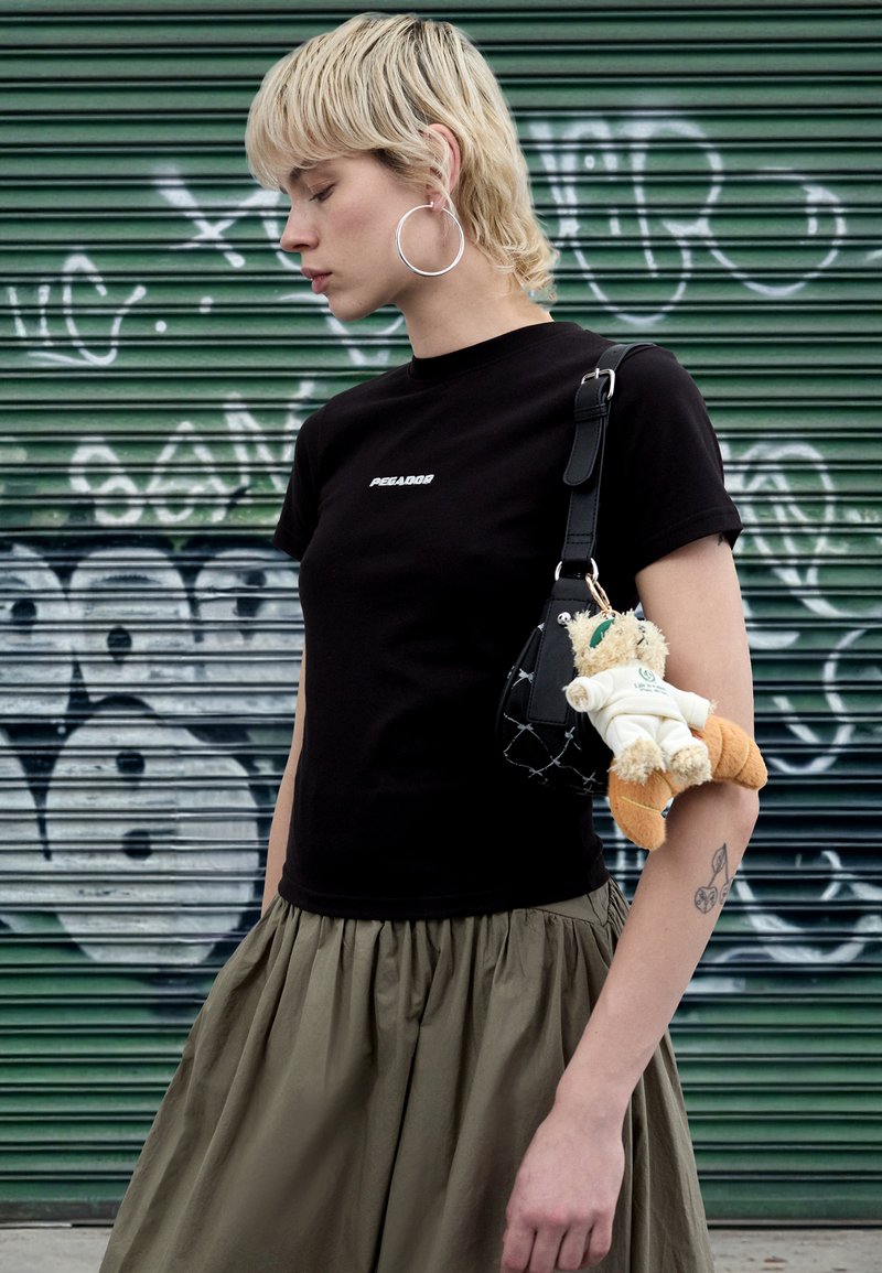 Camiseta preta com logótipo, saia plissada verde-oliva e uma mala preta com um brinquedo de peluche castanho claro. O fundo mostra graffiti verde e branco.