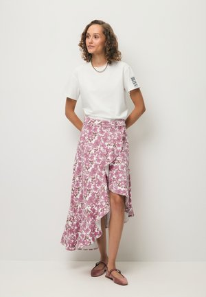 Vrouw met krullend haar, die een wit shirt, een roze asymmetrische bloemenrok en roze platte schoenen draagt, staand tegen een effen lichte achtergrond.