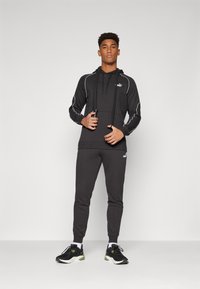 Black athletic hoodie with white accents, paired with matching joggers, featuring a drawstring hood. Worn with black and green running shoes.