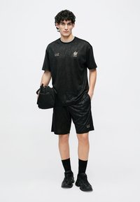 Czarny sportowy strój składający się z koszulki z krótkim rękawem i spodenek z teksturowanym, geometrycznym wzorem, uzupełniony czarnymi trampkami i małą torbą.