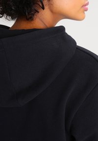 Sweatshirt noir à capuche montrant le tissu texturé et les détails des coutures, avec un accent sur la zone de la capuche et du col, présentant un design minimaliste.