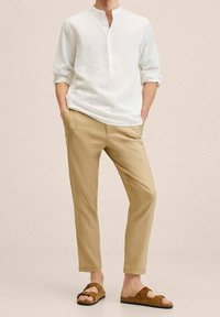 Person, der ein weißes Langarmshirt, beigen Hosen und braunen Sandalen mit offenen Zehen trägt, steht mit den Händen in den Taschen vor einem einfarbigen Hintergrund.