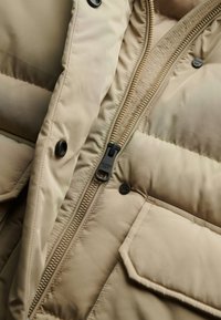 Chaqueta acolchada beige con cierre frontal, botones de presión negros, textura acolchada y un logo visible en la cremallera.