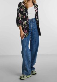 Mujer con vaqueros de pierna ancha azul, camisa blanca, blazer negro con estampado floral y zapatillas blancas con detalles verdes, de pie frente a un fondo liso.