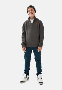 Grå strikket trøje med halvlynlås og læderlogo-patch, kombineret med mørkegrønne cargo-bukser og hvide sneakers med sorte detaljer.