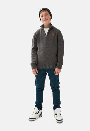 Grå strikket trøje med halvlynlås og læderlogo-patch, kombineret med mørkegrønne cargo-bukser og hvide sneakers med sorte detaljer.