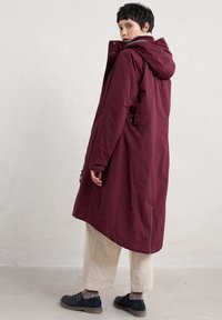 Lange bordeauxrode jas met een capuchon, rits aan de voorkant en zijzakken. Heeft een gladde textuur en een licht oversized ontwerp. Ggedragen met crème kleurige corduroy broeken.