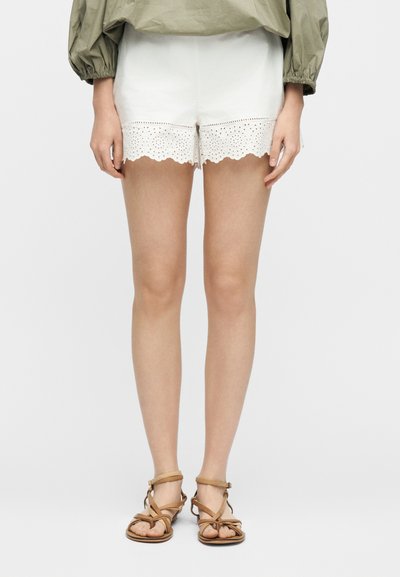 Γυναίκα που φοράει λευκά σορτς με τελείωμα από δαντέλα eyelet, πράσινο-ελαιί πράσινο μακρυμάνικο μπλουζάκι και καφέ ίσια σανδάλια με λουράκια, στέκεται μπροστά σε απλό φόντο.