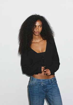 Femme aux longs cheveux bouclés portant un cardigan noir court par-dessus un haut noir et un jean bleu ample, debout devant un fond blanc uni.