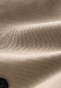 Material bej cu țesătură fină și texturată; prezintă modele subtile diagonale și un nasture negru vizibil la margine.