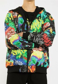 Personne portant un hoodie zippé multicolore de style graffiti avec des motifs abstraits et des tags, les bras croisés sur une chemise noire et un pantalon.
