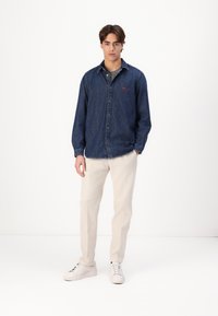 Denim shirt in dark blue with a button-up front and long sleeves, featuring a small red logo. Paired with light-colored pants and white sneakers.