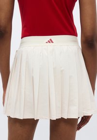 Κρεμ πλισέ φούστα με αντίθετη μέση, διακοσμημένη με μικρό κόκκινο λογότυπο. Ελαφρύ ύφασμα με ομαλή υφή.