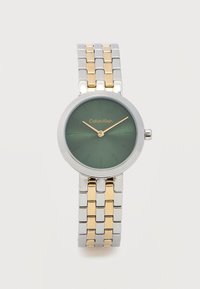 Ρολόι χειρός Calvin Klein με πράσινο καντράν, χρυσά δείκτες, ασημένιο και χρυσό μπρασελέ με συνδέσμους, και μινιμαλιστικούς δείκτες ώρας σε λευκό φόντο.