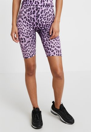 Persona che indossa pantaloni biker a stampa leopardata viola e scarpe da ginnastica nere su uno sfondo bianco semplice.