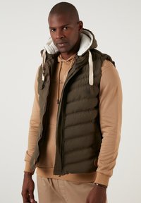 Olive green padded vest with a hood and cream lining, featuring a front zip, toggle drawstring, and snap buttons on the side.