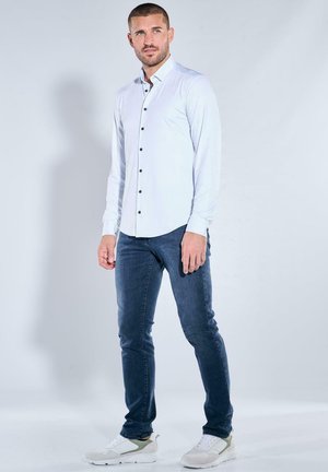 Mann steht, trägt ein weißes Hemd mit Knöpfen, dunkelblaue Jeans und weiße Sneaker vor einem einfarbigen hellen Hintergrund.