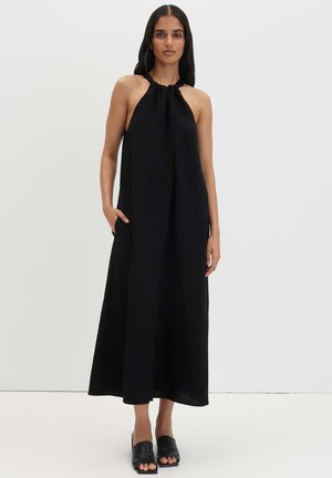 Frau mit langen dunklen Haaren, die ein ärmelloses schwarzes Neckholderkleid und schwarze offene Absatz-Mules trägt, steht vor einem schlichten Hintergrund.