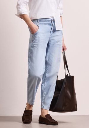 Persona con vaqueros azul claro ajustados, camisa blanca, mocasines marrones, con una mano en el bolsillo y sosteniendo un gran bolso tote negro.