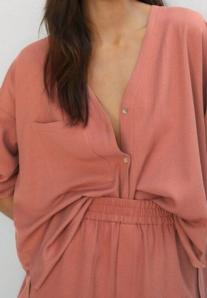 Femme portant un chemisier ample couleur rose poussiéreux avec boutons et poche poitrine, rentrée dans un pantalon assorti à taille élastique.