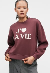 Burgundy sweatshirt with a crew neckline and dropped shoulders. Features white "J'♥ LA VIE" text on the front. Soft fabric.