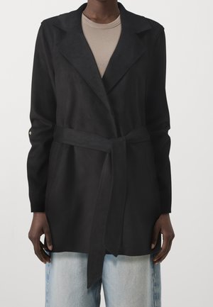 Personne portant une veste noire ceinturée sur un haut beige et un jean bleu clair, debout devant un fond blanc uni.