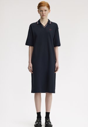 Persona joven con cabello rojo que lleva un vestido tipo polo azul marino hasta la rodilla y mocasines negros con calcetas negras, de pie sobre un fondo liso.