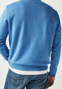 Sweatshirt bleu en tissu doux, avec poignets et ourlet côtelés, au design simple. Porté sur une chemise blanche et associé à un jean en denim.