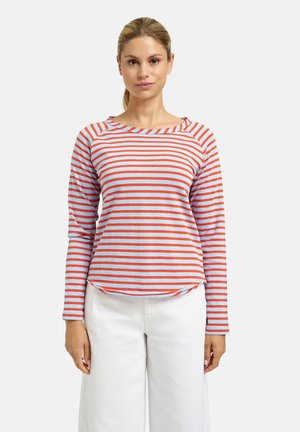 Frau mit blonden Haaren im Pferdeschwanz, die ein blau-orange gestreiftes Langarmshirt und eine weiße Hose vor weißem Hintergrund trägt.