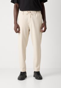 Un pantalon beige à taille élastique présente une fermeture à cordon, des poches latérales et une coupe décontractée, associé à des baskets noires.