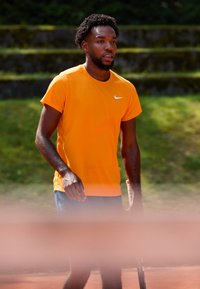 Lys orange atletisk skjorte med korte ærmer, forsynet med et lille hvidt Nike-logo. Båret med blå shorts på en tennisbane.
