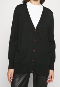 Mustat neuletakki, jossa on V-pääntie, napitus edessä ja kaksi etupussitaskua. Valmistettu pehmeästä, kevyestä materiaalista, jossa on ribattu hihansuut.