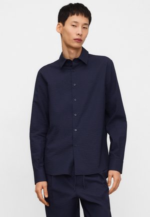 Młody mężczyzna ubrany w granatową koszulę z długim rękawem z teksturowanego materiału oraz dopasowane spodnie, na tle jednolitego jasnego tła.