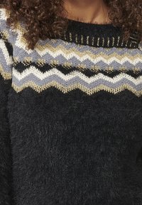 Pull tricoté avec un col rond, base gris foncé, motifs en zigzag en gris clair, blanc et fils dorés. Texture douce.