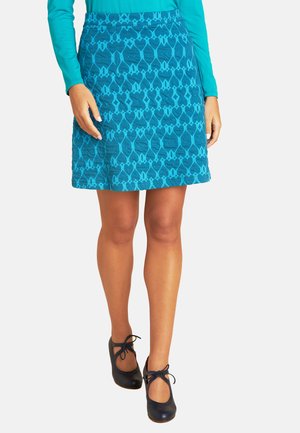 Türkiser Rock mit strukturiertem geometrischen Muster, knielanger Schnitt, kombiniert mit marineblauen Schuhen mit einer Schleifendetail und Ausschnitten.