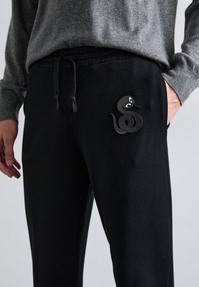 Schwarze Jogginghose mit elastischem Bund, mit einer strukturierten schwarzen Leder-Schlangenapplikation am linken Oberschenkel. Der Stoff ist weich und glatt.