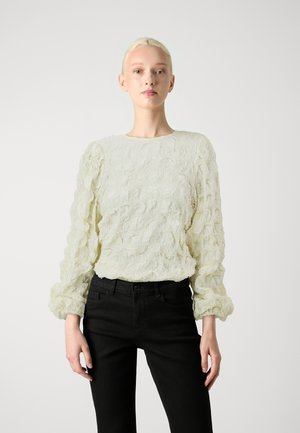 Lys cremet bluse med struktureret, flæset stof og lange pufærmer, parret med sorte tætsiddende jeans. Enkel rund halsudskæring.