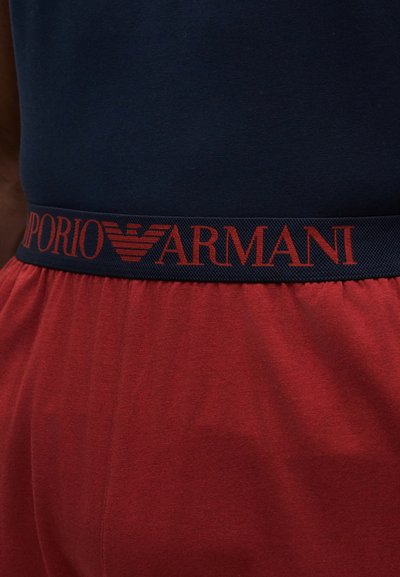 Ελαστική μέση με κόκκινο λογότυπο Emporio Armani σε σκοτεινό μπλε λωρίδα πάνω από φαρδιά κόκκινα παντελόνια ή σορτς.