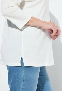 Bílý dlouhý rukáv s bočním rozparkem, v kombinaci s modrými džínovými kalhotami. Měkká textura a neformální design.