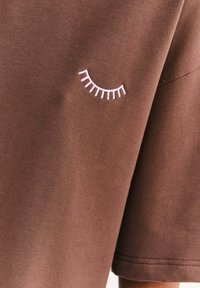 Brązowy bawełniany t-shirt z krótkimi rękawami, okrągłym dekoltem i różowym haftem oka na lewym rękawie.