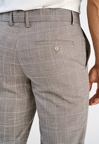 Homme portant un pantalon habillé à carreaux beige clair avec une poche arrière boutonnée et une chemise blanche rentrée.