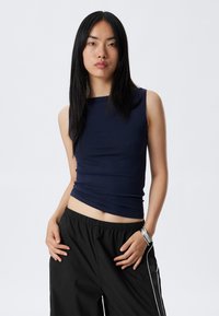 Débardeur sans manches bleu marine en tissu extensible avec un décolleté carré et une coupe ajustée, associé à un pantalon noir ample avec des rayures sur les côtés et une taille élastique.
