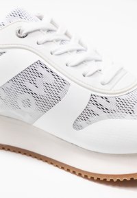 Bílé tenisky s síťovanou textilií, strukturovanými černými detaily a gumovou podrážkou. Mají kulaté tkaničky a hladký, zakřivený design svršku.