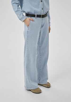 Asmuo dėvi šviesiai mėlynas platėjančias džinsines kelnes, atitinkančią džinsinę marškinių dalį, juodą diržą ir smėlio spalvos slip-on batus, stovintis su viena ranka kišenėje.