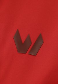 Des formes géométriques plates de couleur marron ressemblant à des chevrons, placées sur un fond en tissu texturé rouge. Un design angulaire proéminent avec une surface lisse.