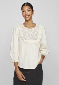 Blouse couleur crème avec un col volanté ornée de broderies florales. Manches longues avec des poignets élastiques et des détails décoratifs à l'ourlet.