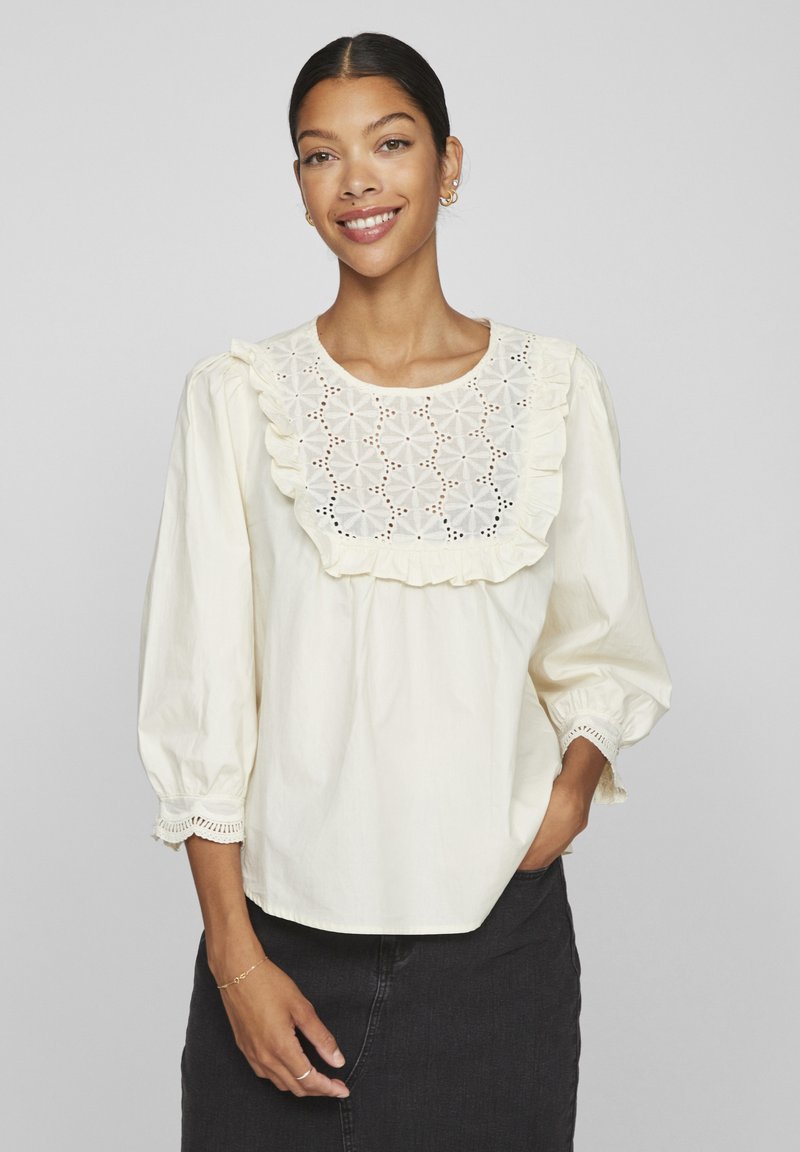 Blouse couleur crème avec un col volanté ornée de broderies florales. Manches longues avec des poignets élastiques et des détails décoratifs à l'ourlet.